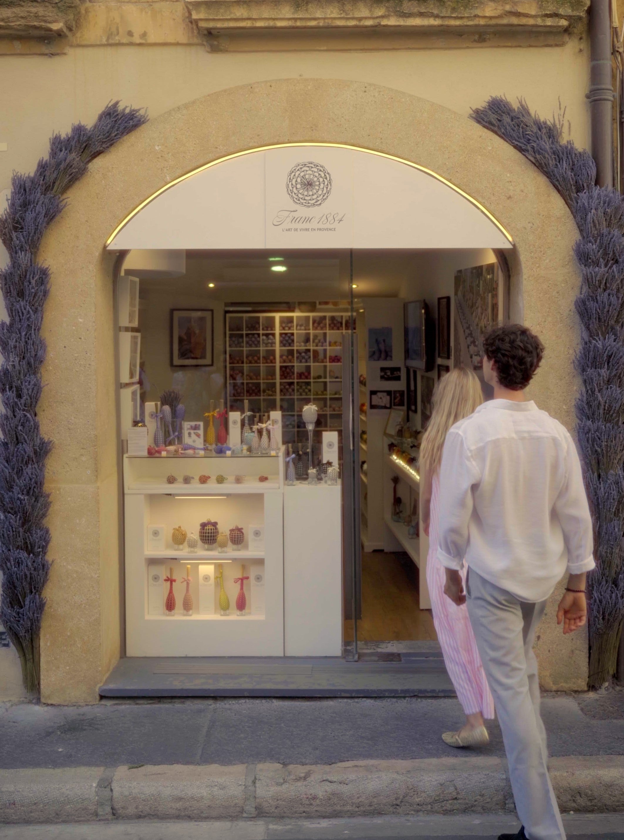 Vue extérieure de la boutique Franc 1884 à Aix-en-Provence. Un couple entre sous l'arche décorée de lavande, devant une vitrine présentant les fuseaux de lavande artisanaux.