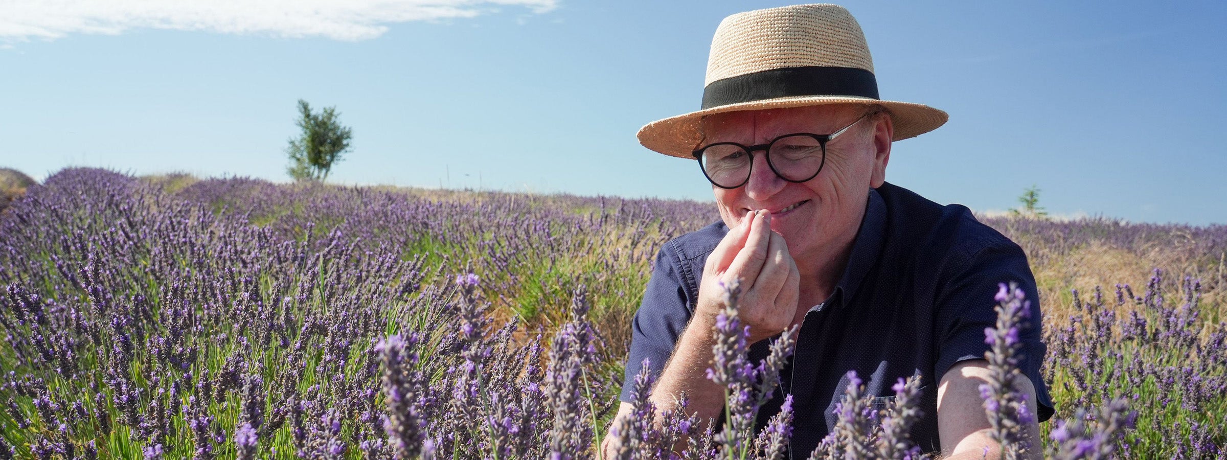 Portrait de Philippe Franc souriant, sentant les fleurs de lavande de Provence dans un champ ensoleillé, portant un chapeau de paille.