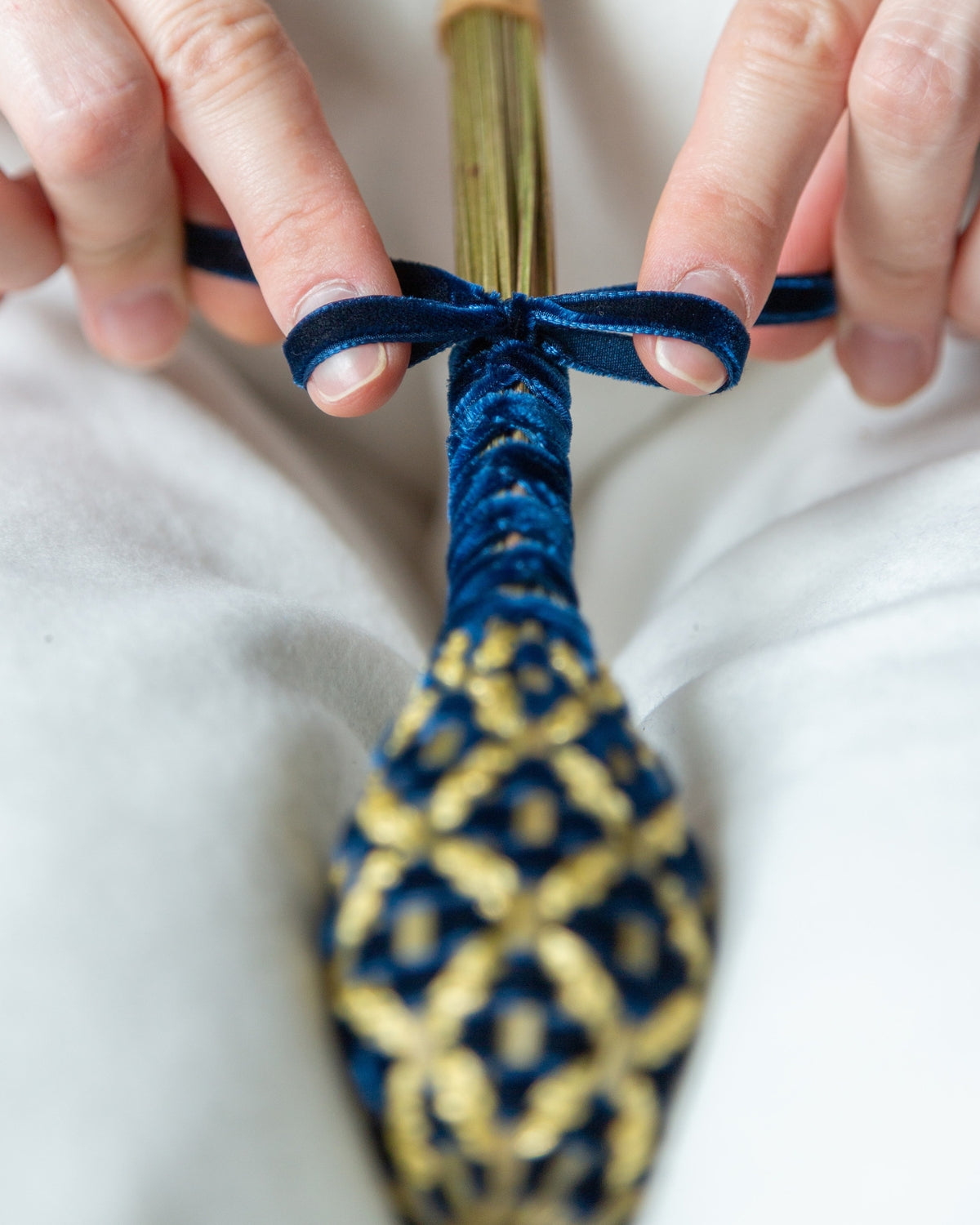 Mains réalisant le nœud de finition en velours bleu marine sur la tige d'un fuseau de lavande, témoignant de la délicatesse de l'artisanat français.
